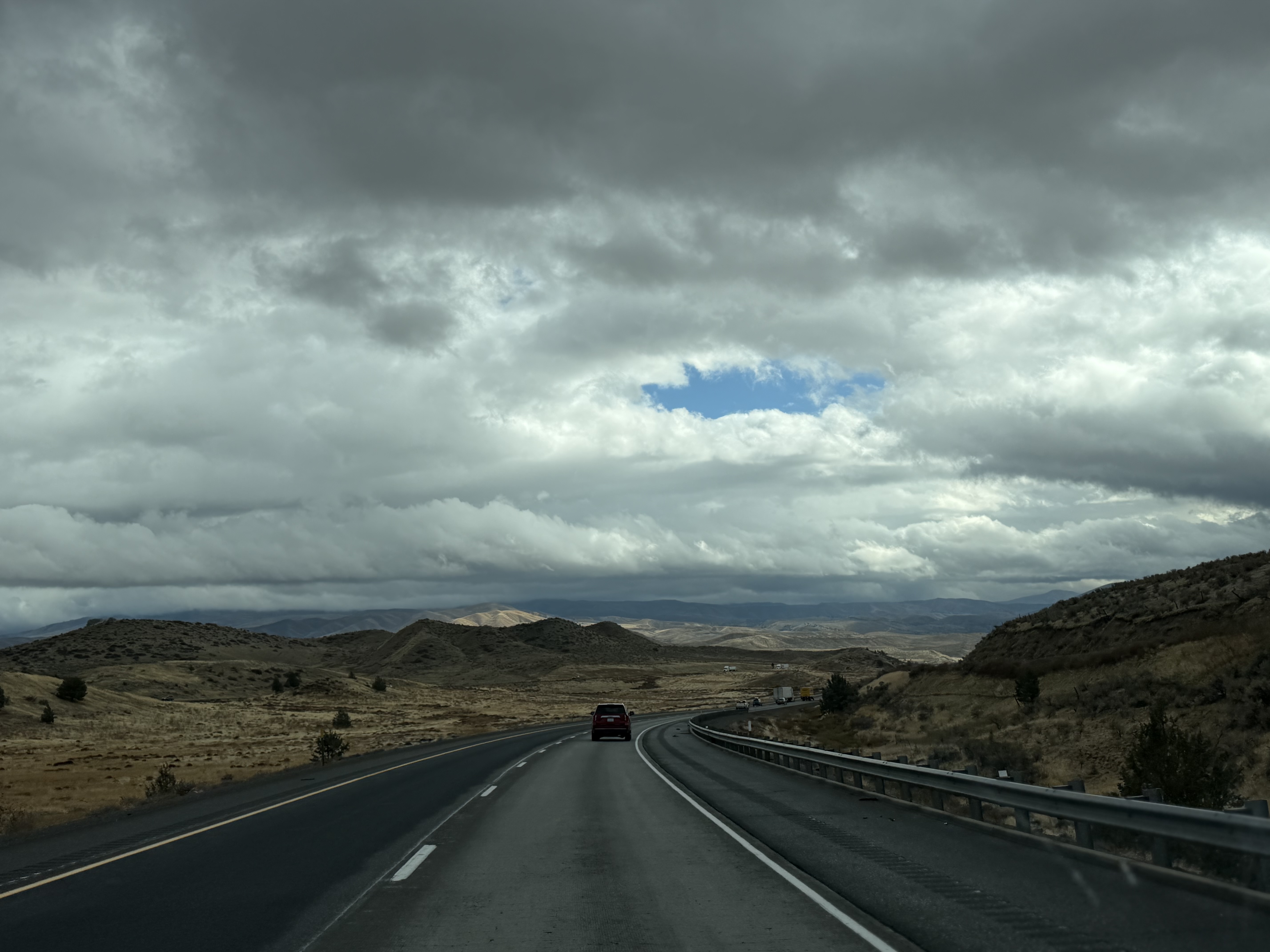 A two-lane highway through dry rolling hills in southern Idaho, broken clouds overhead, a hint of blue sky in the middle.