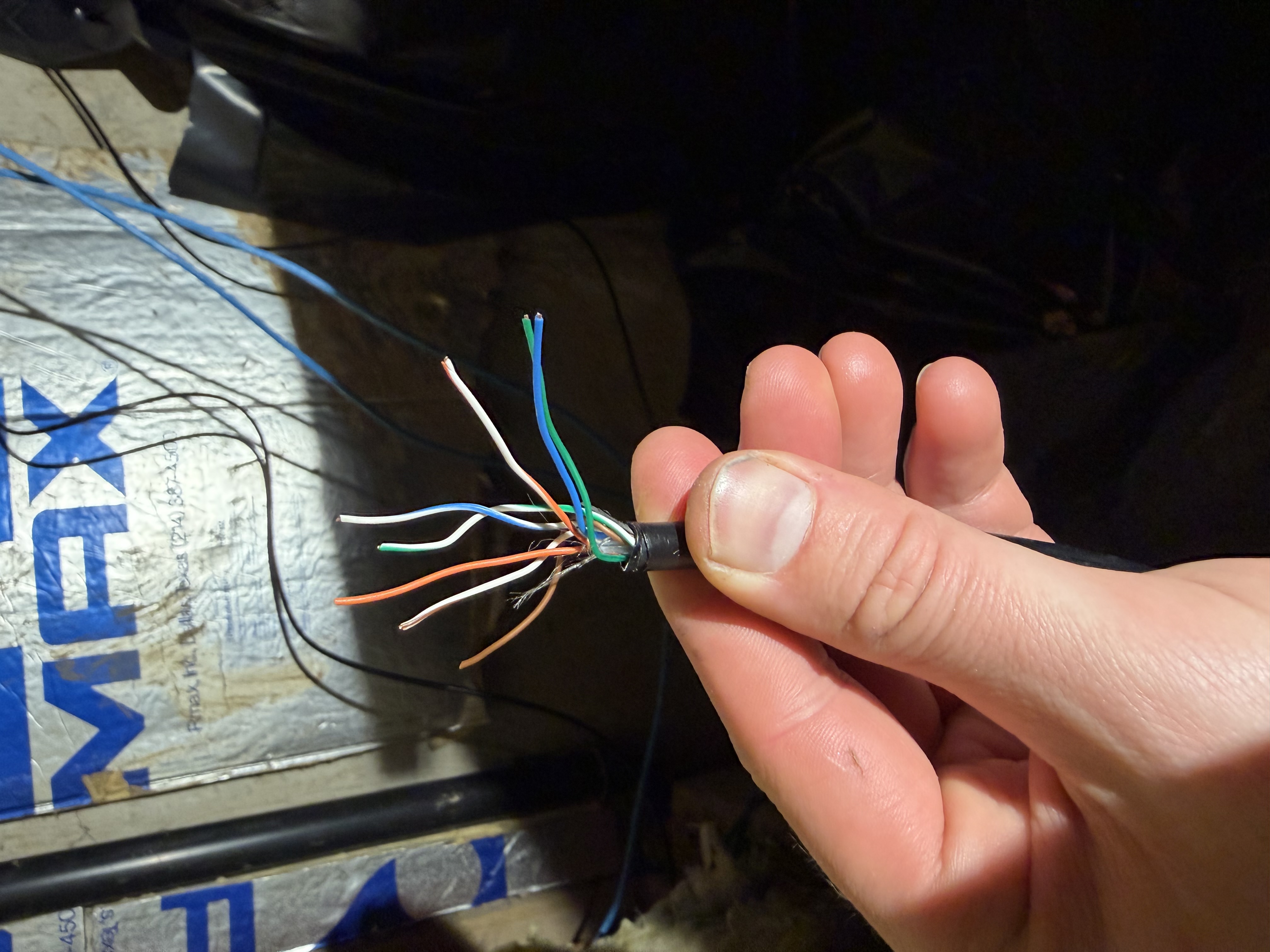 A hand holding the end of a stripped CAT6A cable, eight individual wires fanned out in different colors against a basement insulation backdrop.