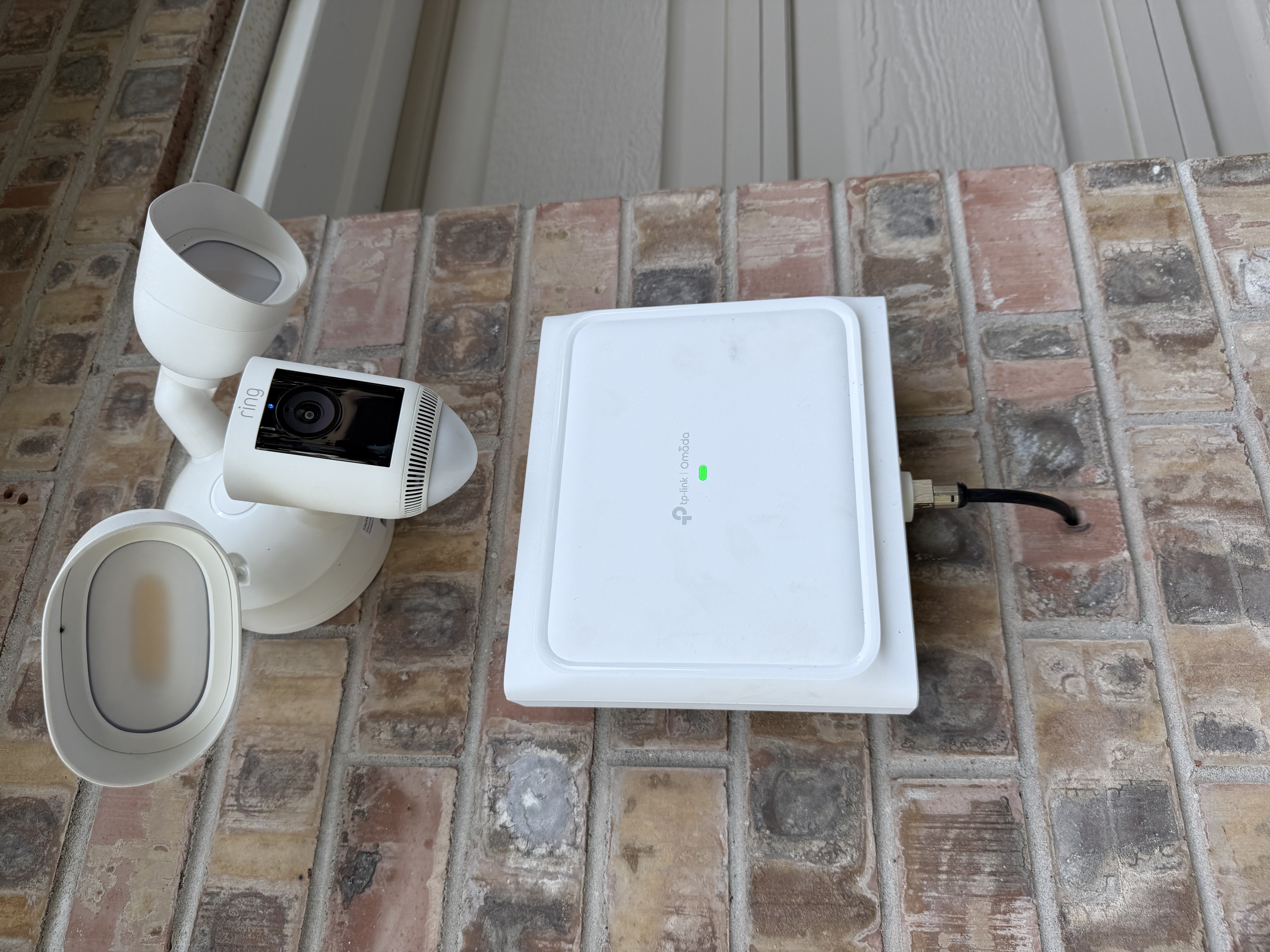 A wall-mounted white Omada outdoor access point bolted to a brick wall, with a Ring spotlight camera and floodlight above it.