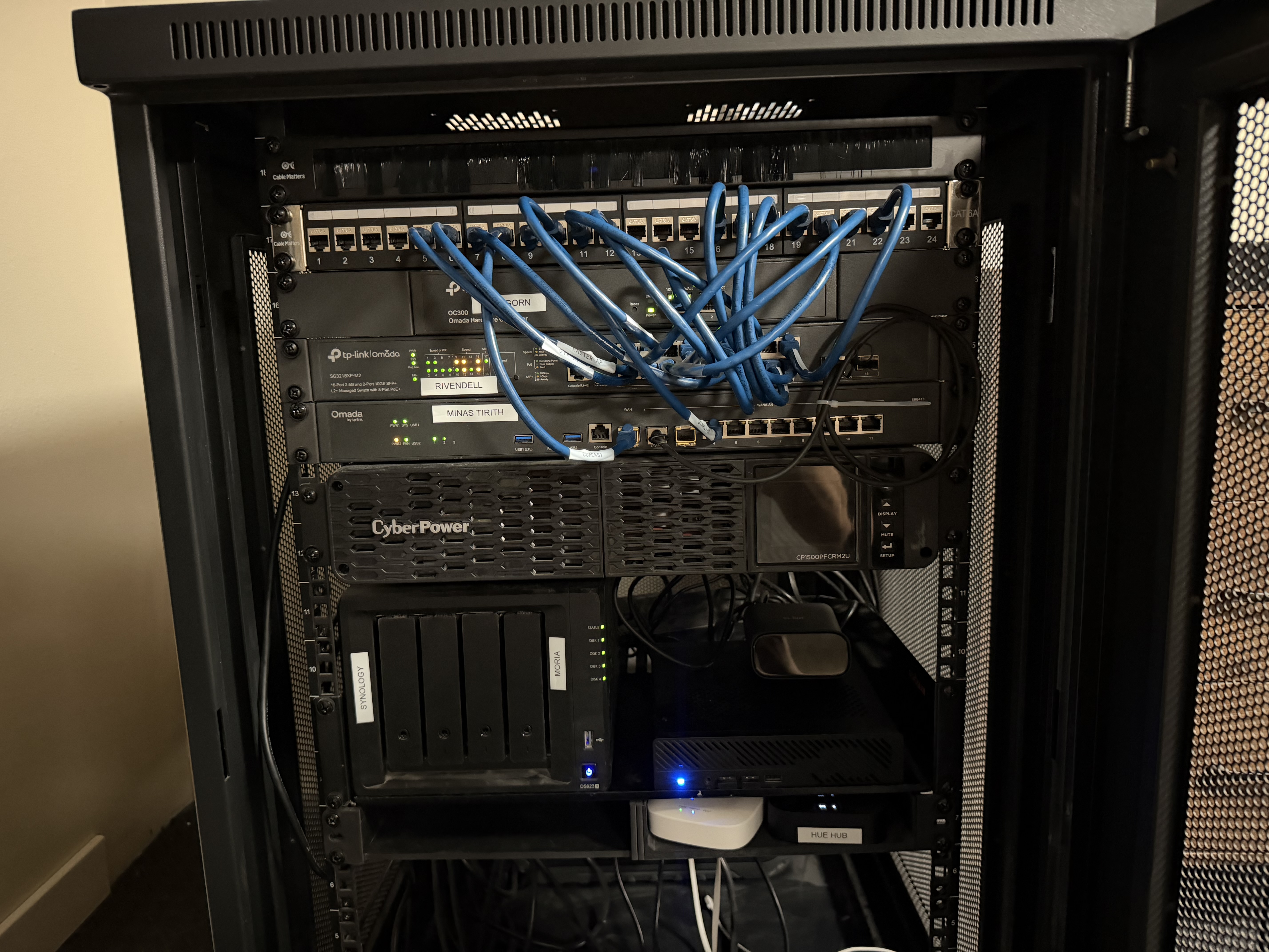 Inside of a black server rack with a Cable Matters CAT6A patch panel at the top, blue patch cables looping down to a TP-Link Omada switch labeled RIVENDELL, an Omada gateway labeled MINAS TIRITH, a CyberPower UPS, a Synology NAS labeled MORIA, and a Hue Hub at the bottom.