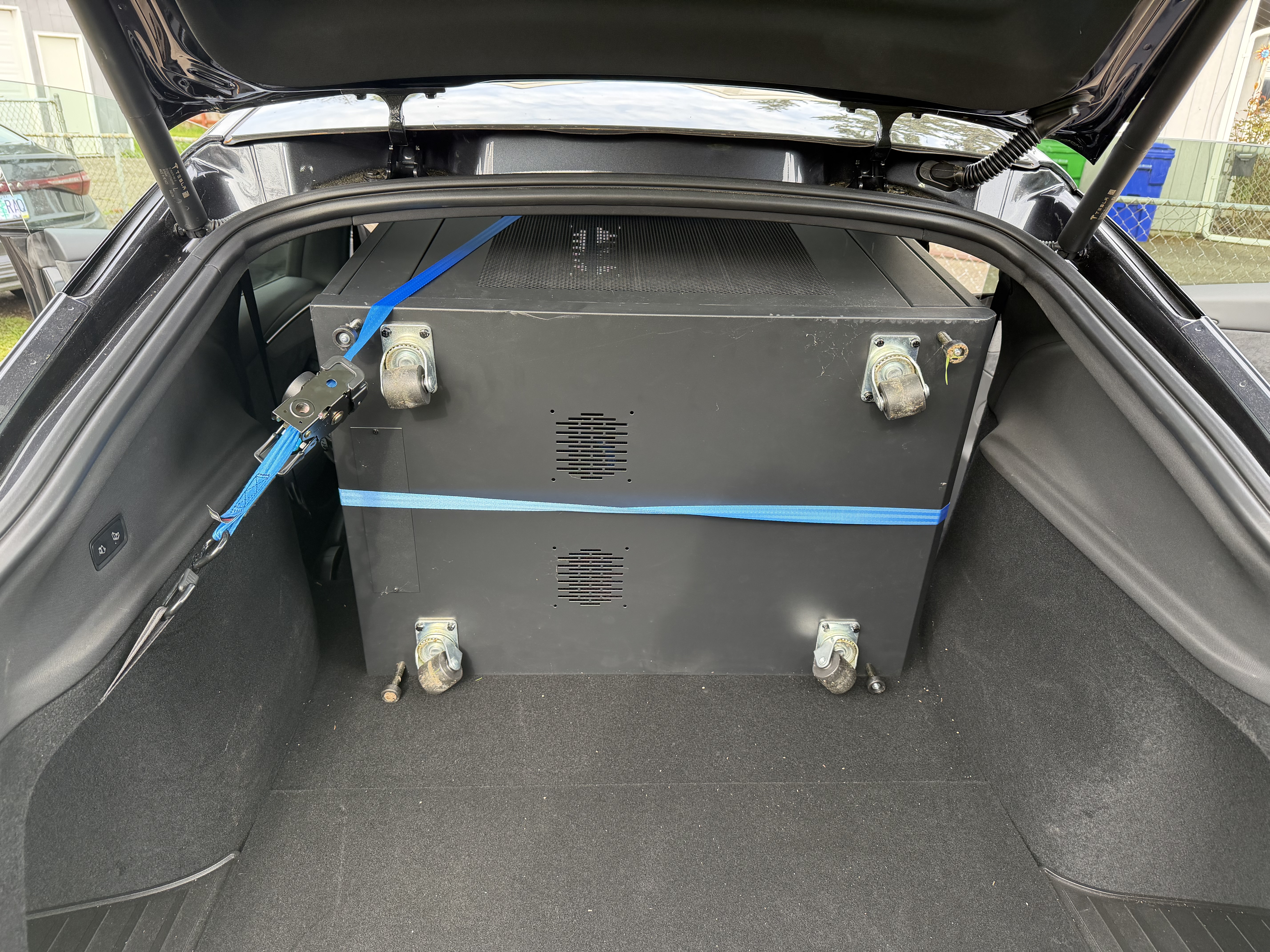A black Startech server rack on casters, strapped down with a blue ratchet tie inside the trunk of a car.