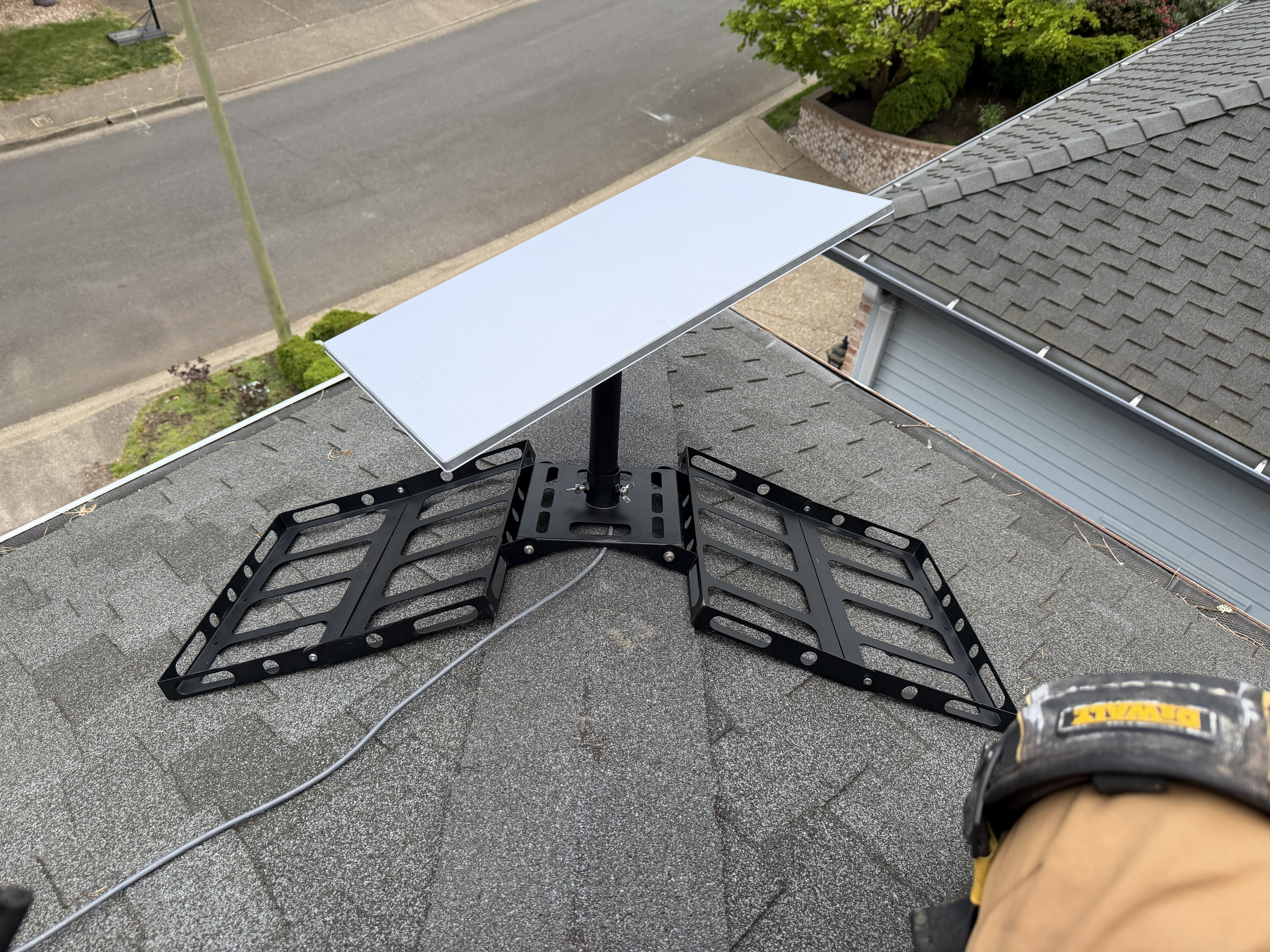 A white Starlink dish mounted to a black non-penetrating roof bracket sitting on dark composition shingles, with the street and houses visible below.