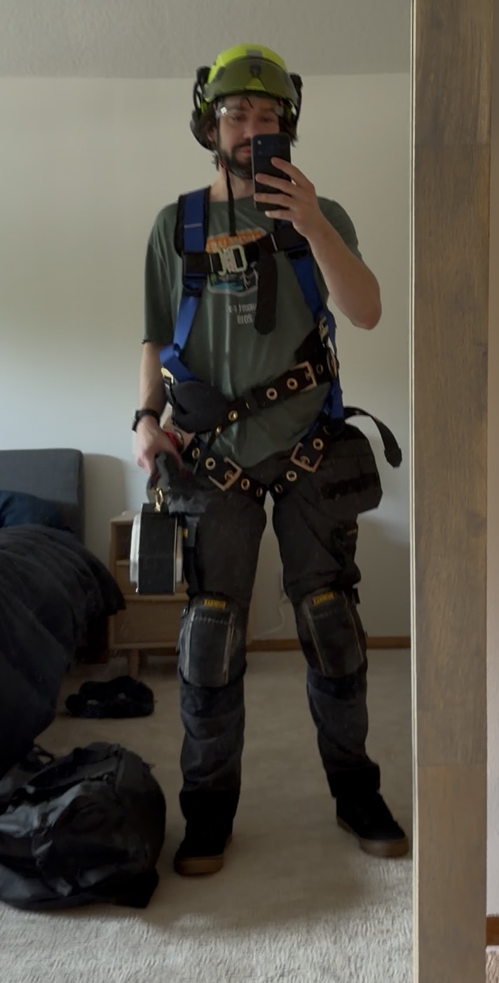 Me in roofing gear — hard hat, safety glasses, fall harness, kneepads — holding a fish tape, taking a mirror selfie before going up to mount the Starlink dish.