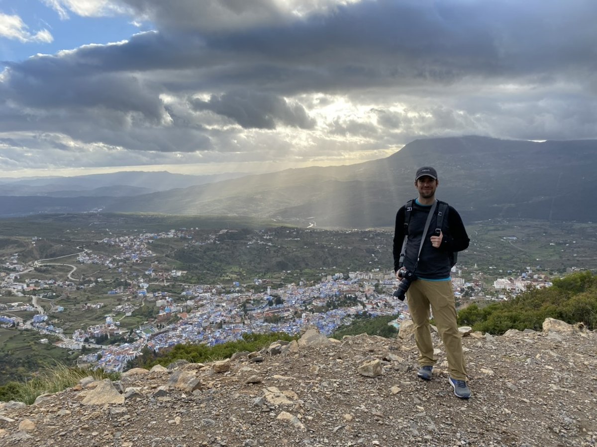 David standing on a ridge above Chefchaouen, Morocco, with the blue-painted city and a dramatic sunlit valley below